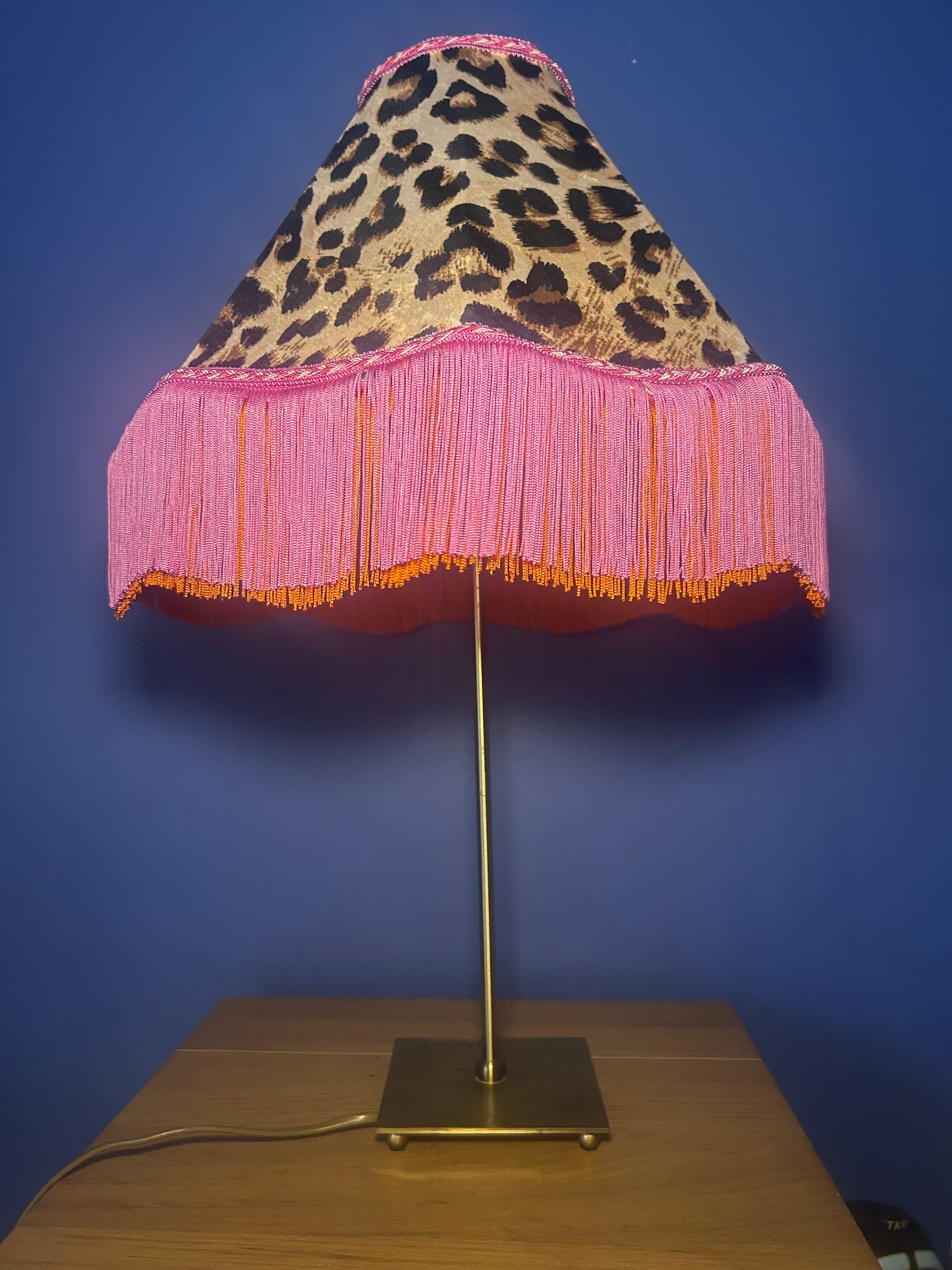 Lamp with leopard print and pink fringe shade on a wooden base against a blue wall.