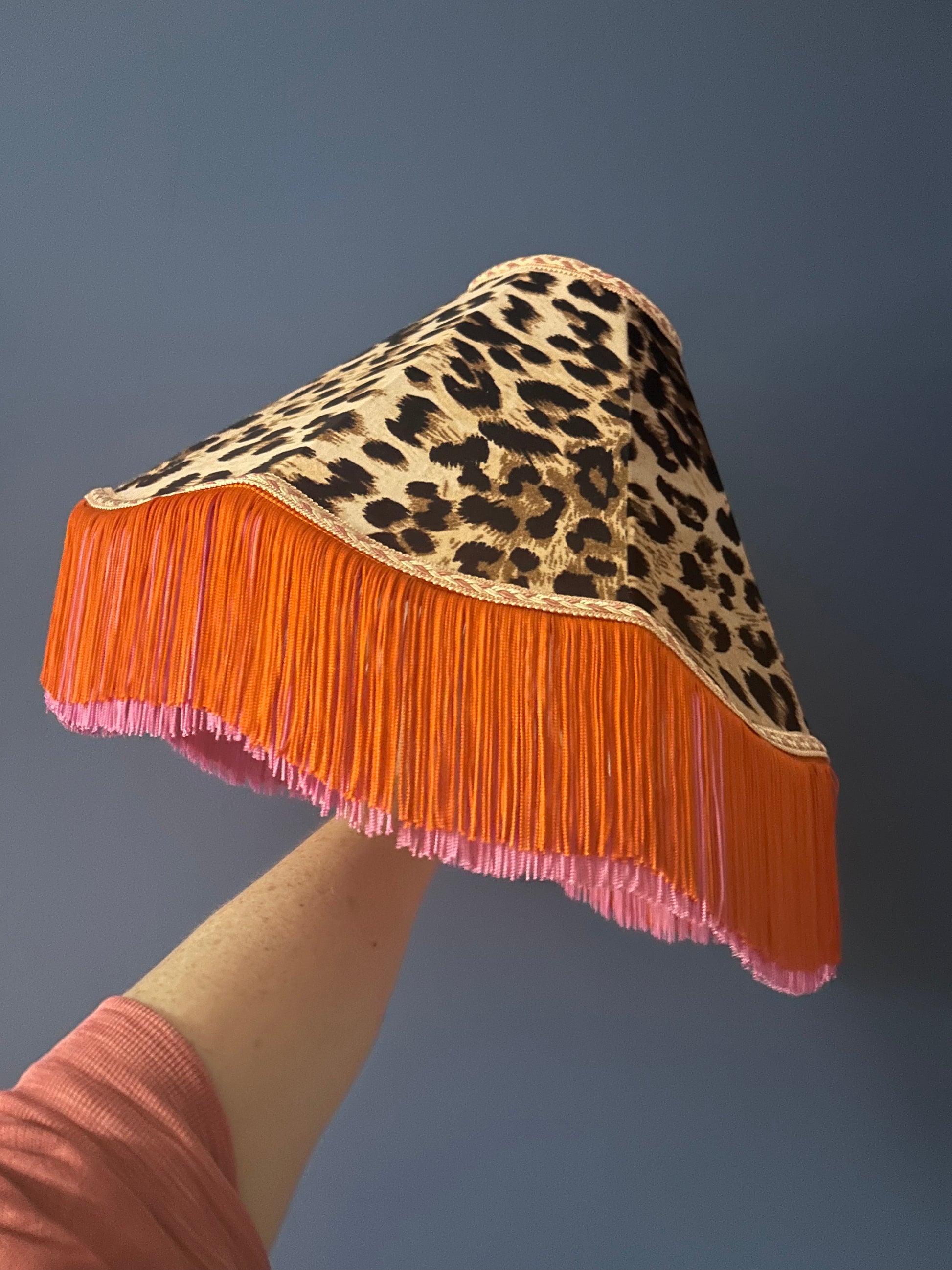 Lampshade with leopard print top and orange fringe bottom held by a hand against a dark background
