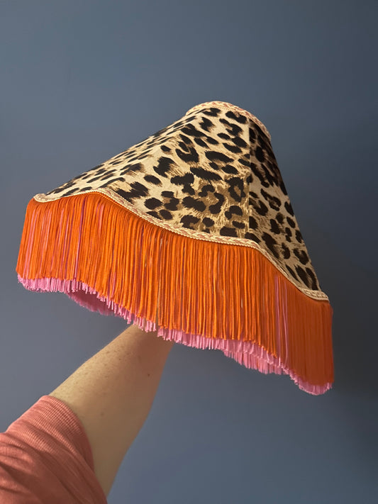 Lampshade with leopard print top and orange fringe bottom held by a hand against a dark background