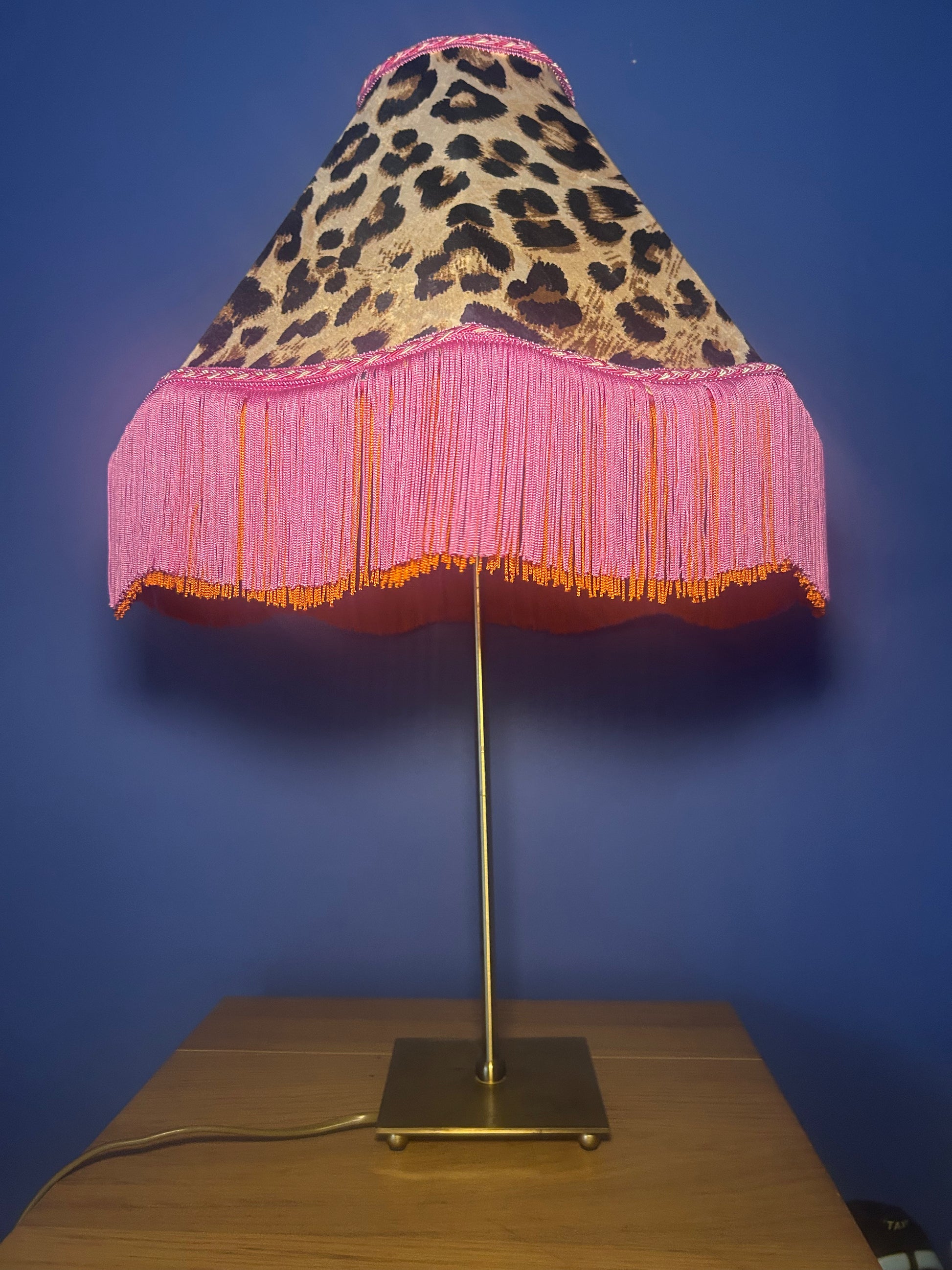 Lamp with leopard print and pink fringe shade on a wooden base against a blue wall.